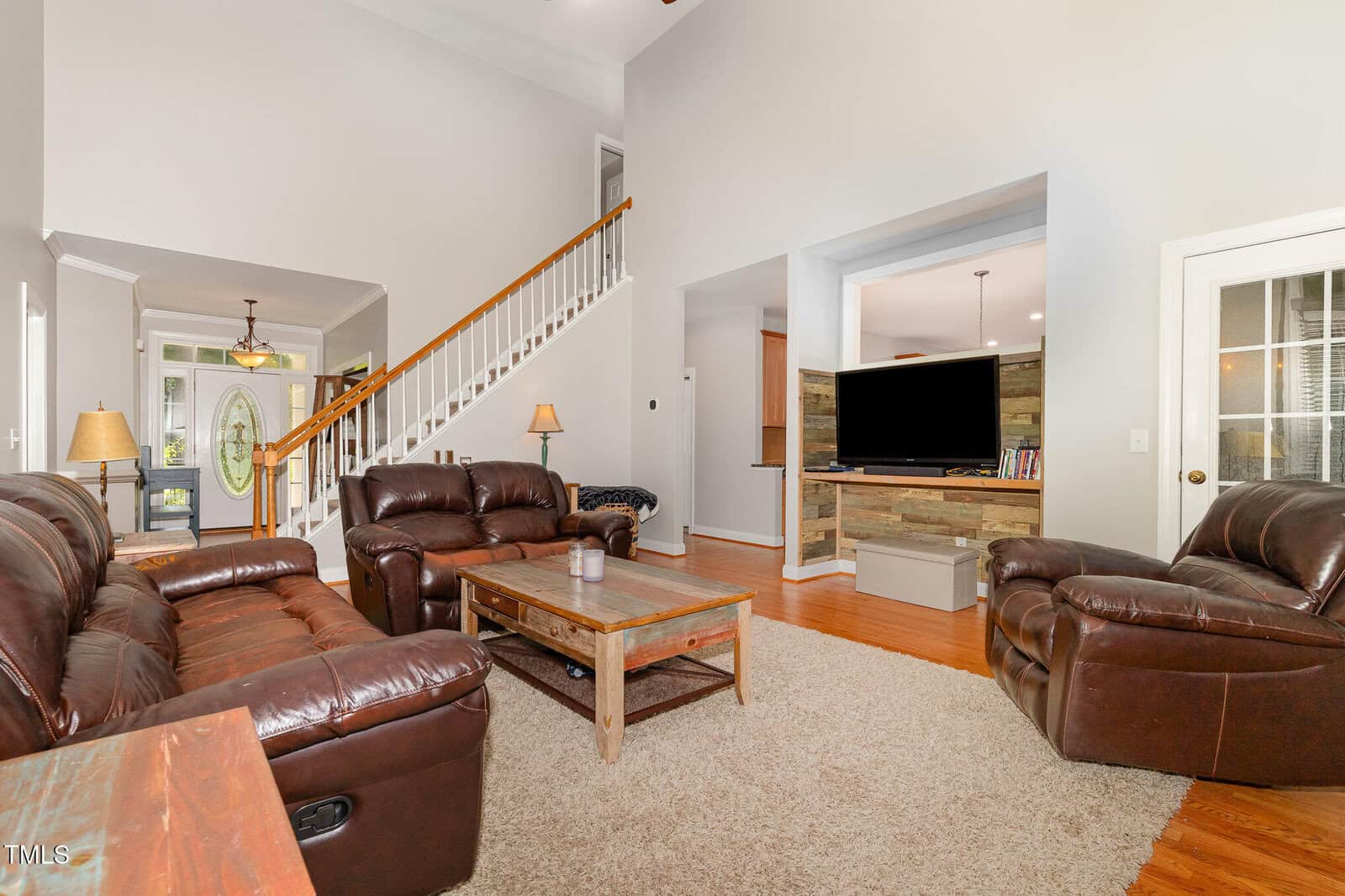A spacious living room with brown leather sofas, a wooden coffee table, a beige rug, and a TV on a stand. A staircase with a wooden handrail leads upstairs, and hardwood floors are visible throughout.