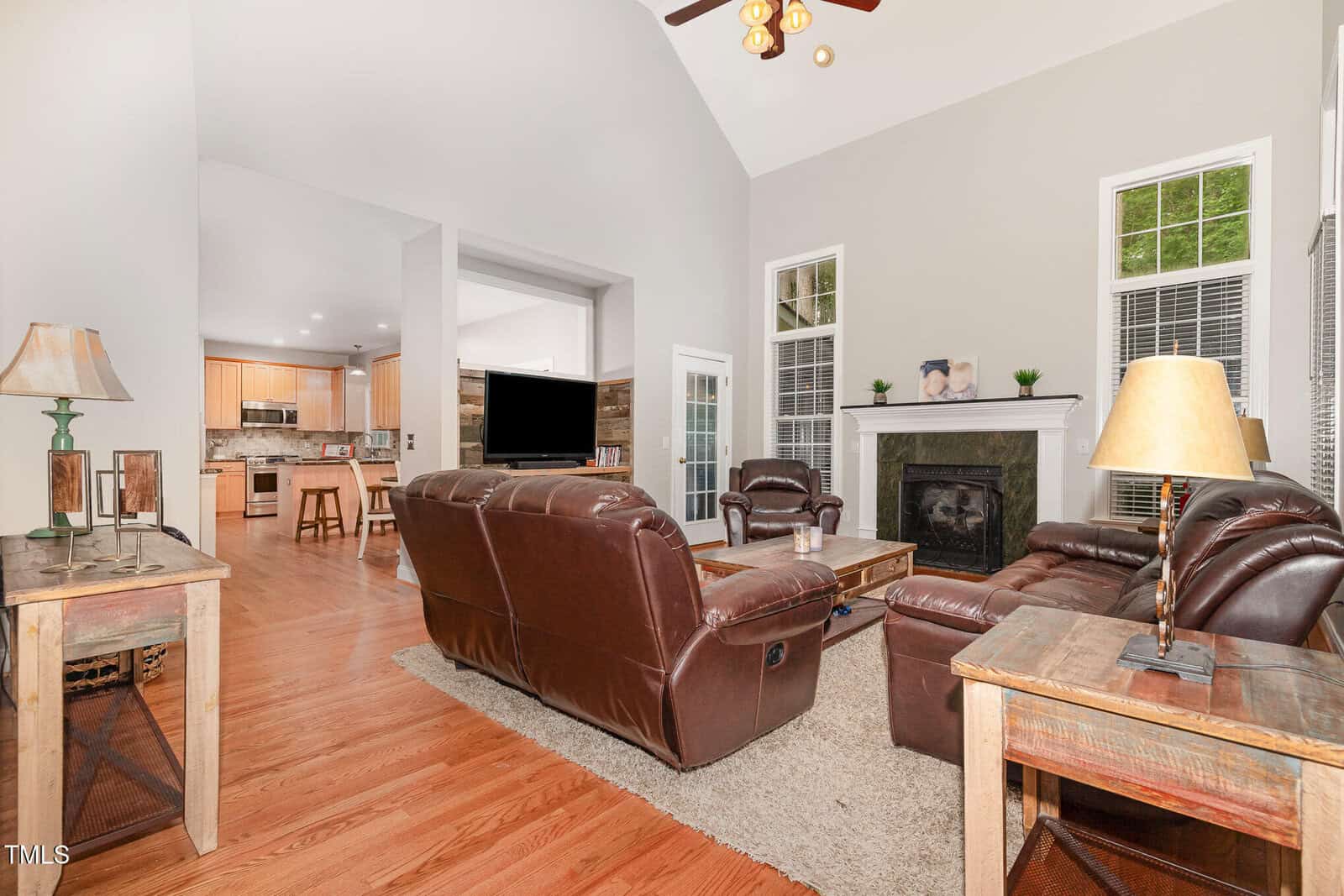 Living room with brown leather sofas, wooden tables, and a fireplace, connected to a kitchen. Hardwood floors and large windows brighten the space. A TV sits on a stand near the fireplace.