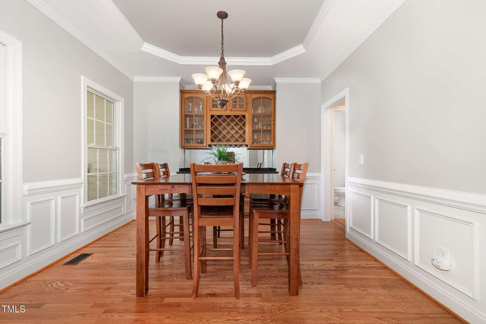 A dining room with hardwood floors, a wooden table and six chairs, a glass-front cabinet, large windows on the left, a chandelier overhead, and white paneled walls with crown molding.