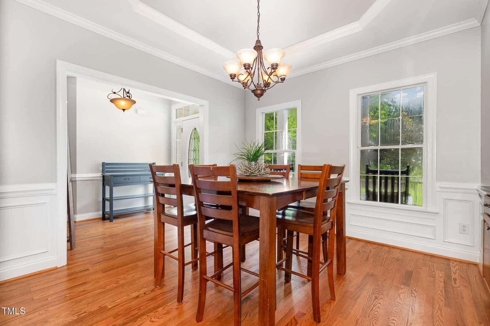 A dining room with a square wooden table and six chairs, hardwood floors, two large windows, light gray walls with white trim, and a chandelier hanging from a recessed ceiling.