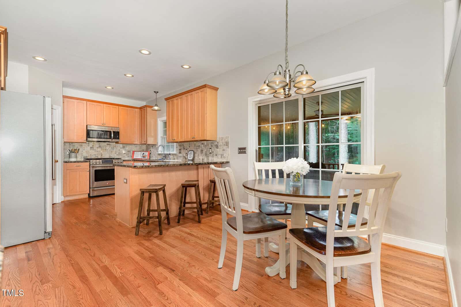 Bright kitchen with wooden floors, light wood cabinets, stainless steel appliances, island with stools, and a round dining table with four chairs by large windows overlooking a covered patio.