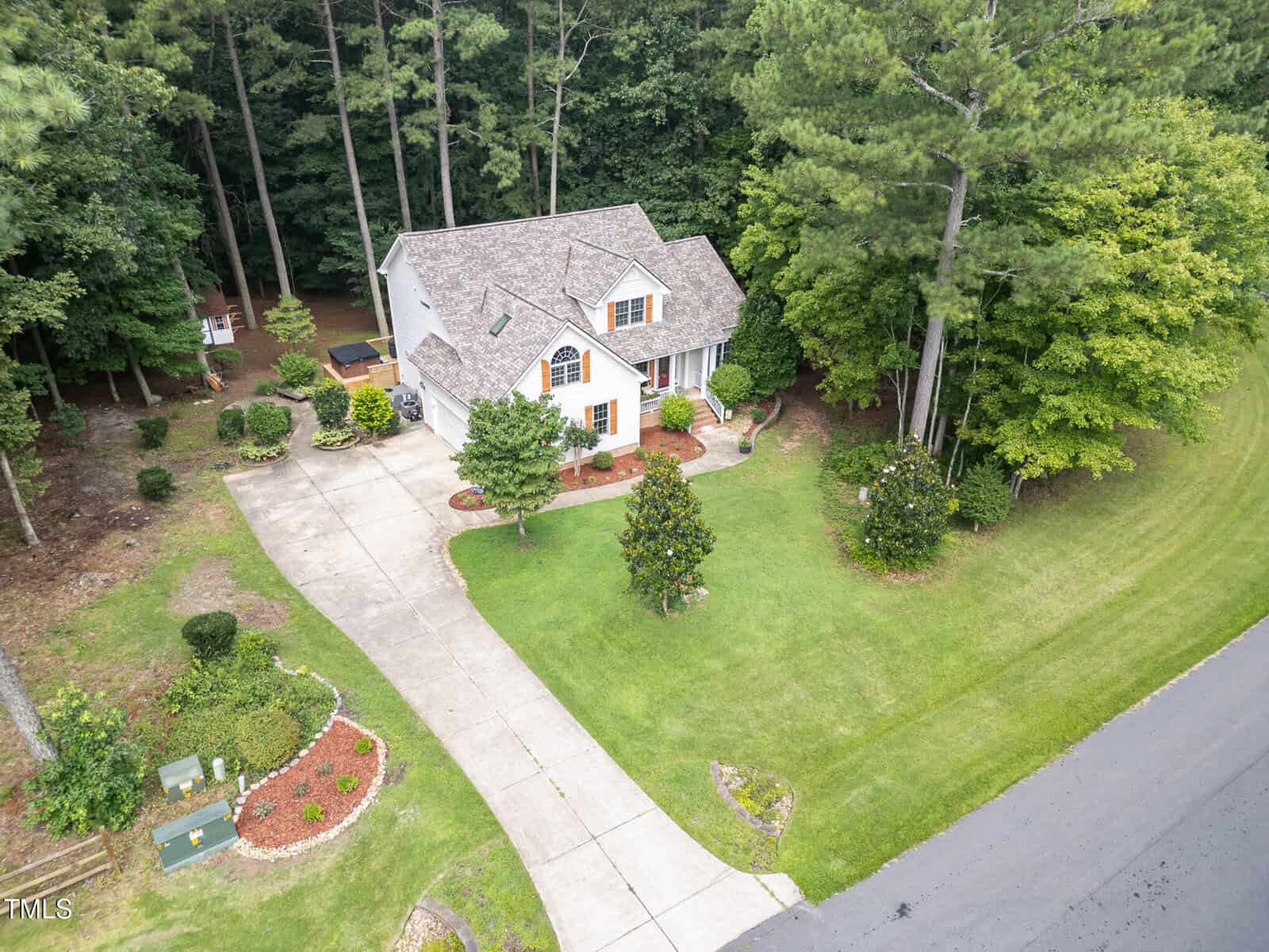 Aerial view of a two-story house with a light-colored exterior, a large driveway, landscaped front yard, and surrounded by tall trees and greenery along a street.
