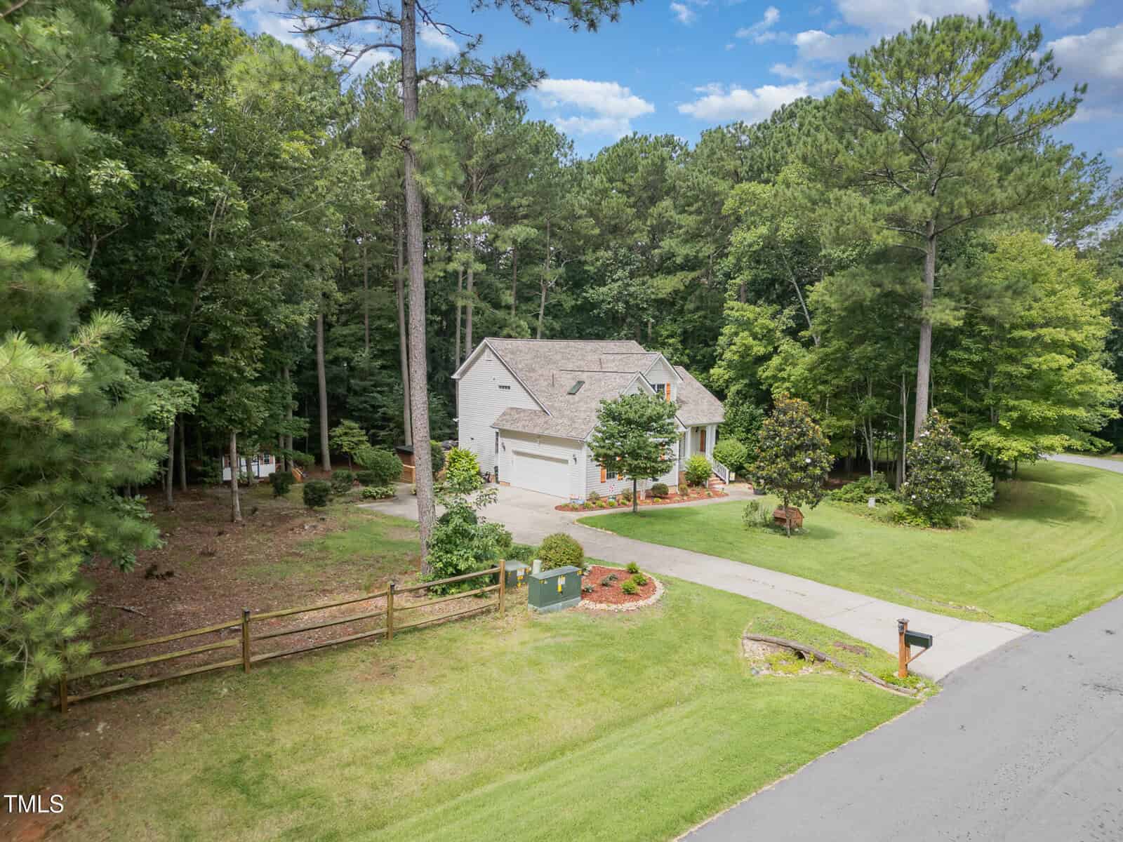 A two-story house with a three-car garage sits on a neatly landscaped lawn, surrounded by tall trees and a wooden fence, adjacent to a paved road on a sunny day.