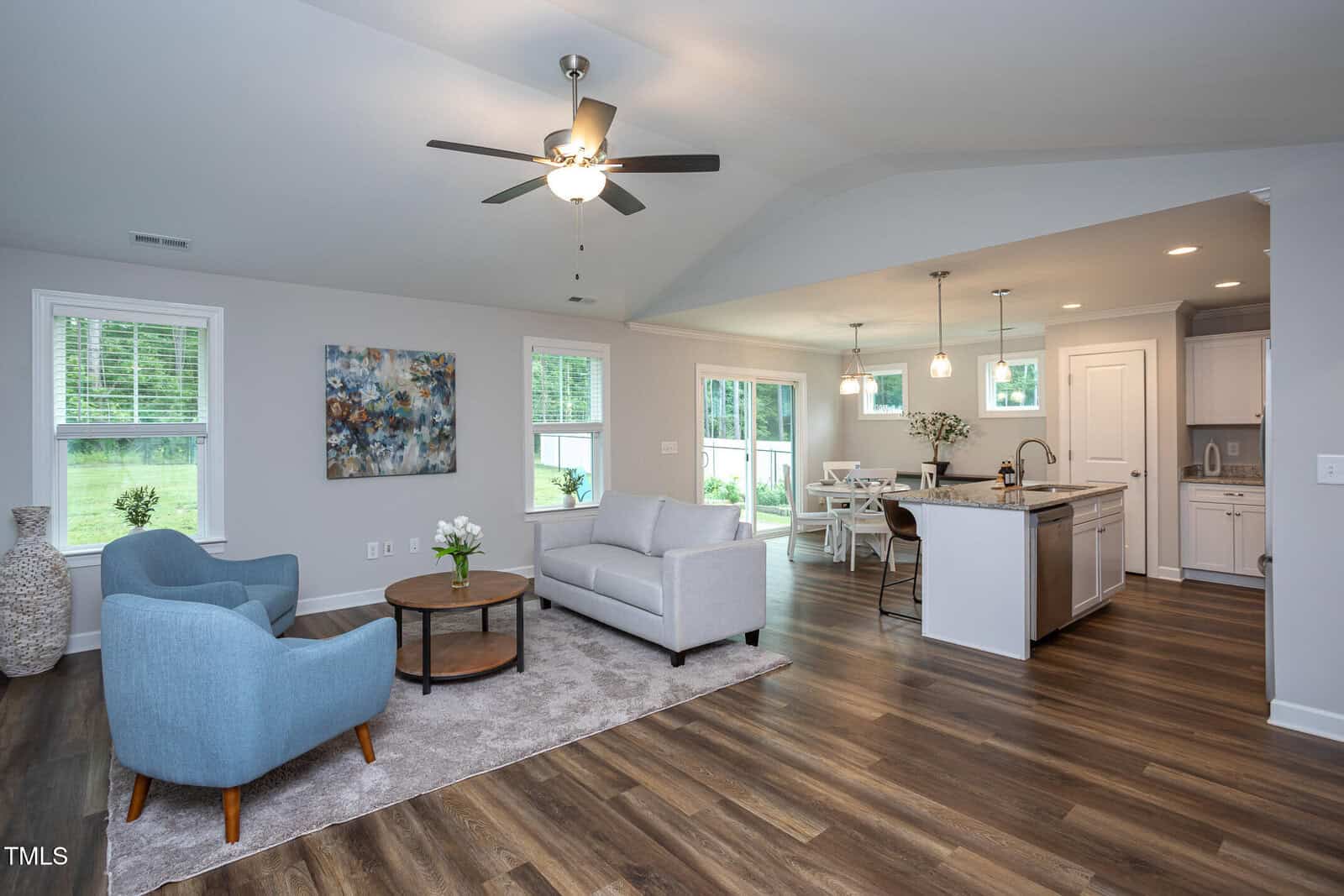 Modern open-concept living and dining area with wood floors, blue armchairs, a light gray sofa, round coffee table, kitchen island, pendant lighting, and large windows letting in natural light.