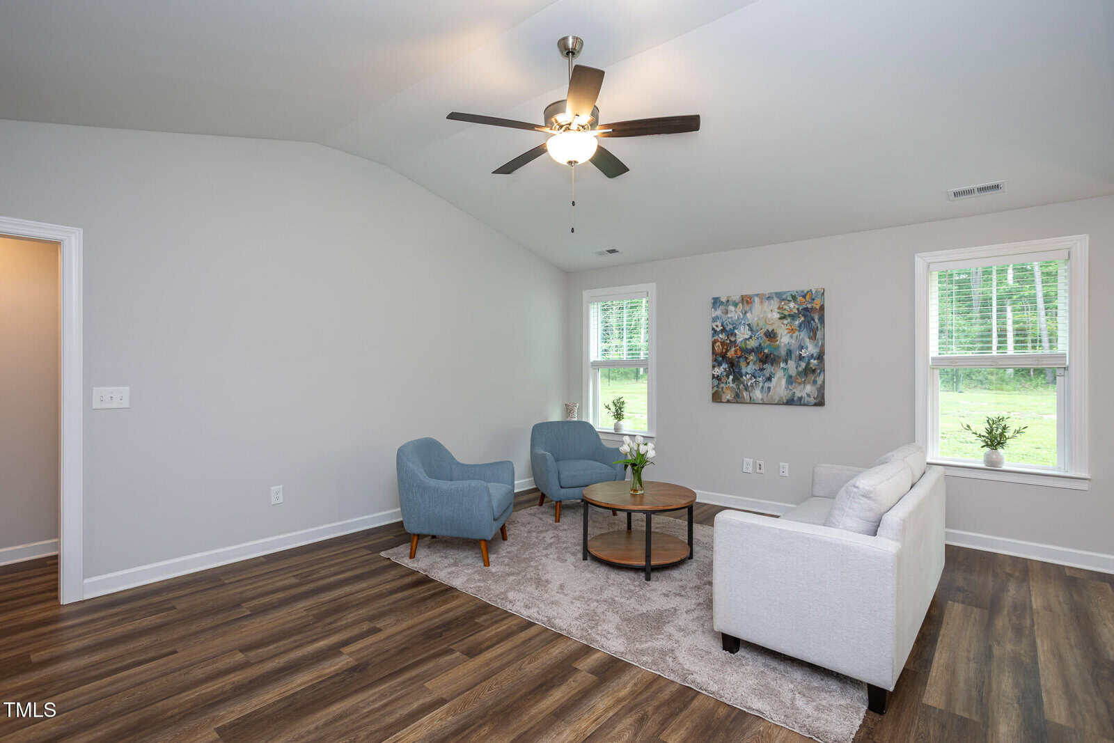 Modern living room with light walls and wood flooring, featuring a white sofa, two blue armchairs, a round coffee table on a gray rug, a ceiling fan with a light, and two windows letting in natural light.