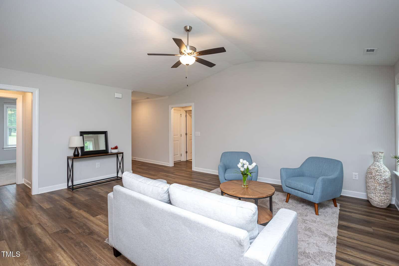 A modern living room with light gray walls, wood flooring, a white sofa, two blue armchairs, a round wooden coffee table with flowers, and a ceiling fan with light. A console table with a mirror is against the back wall.