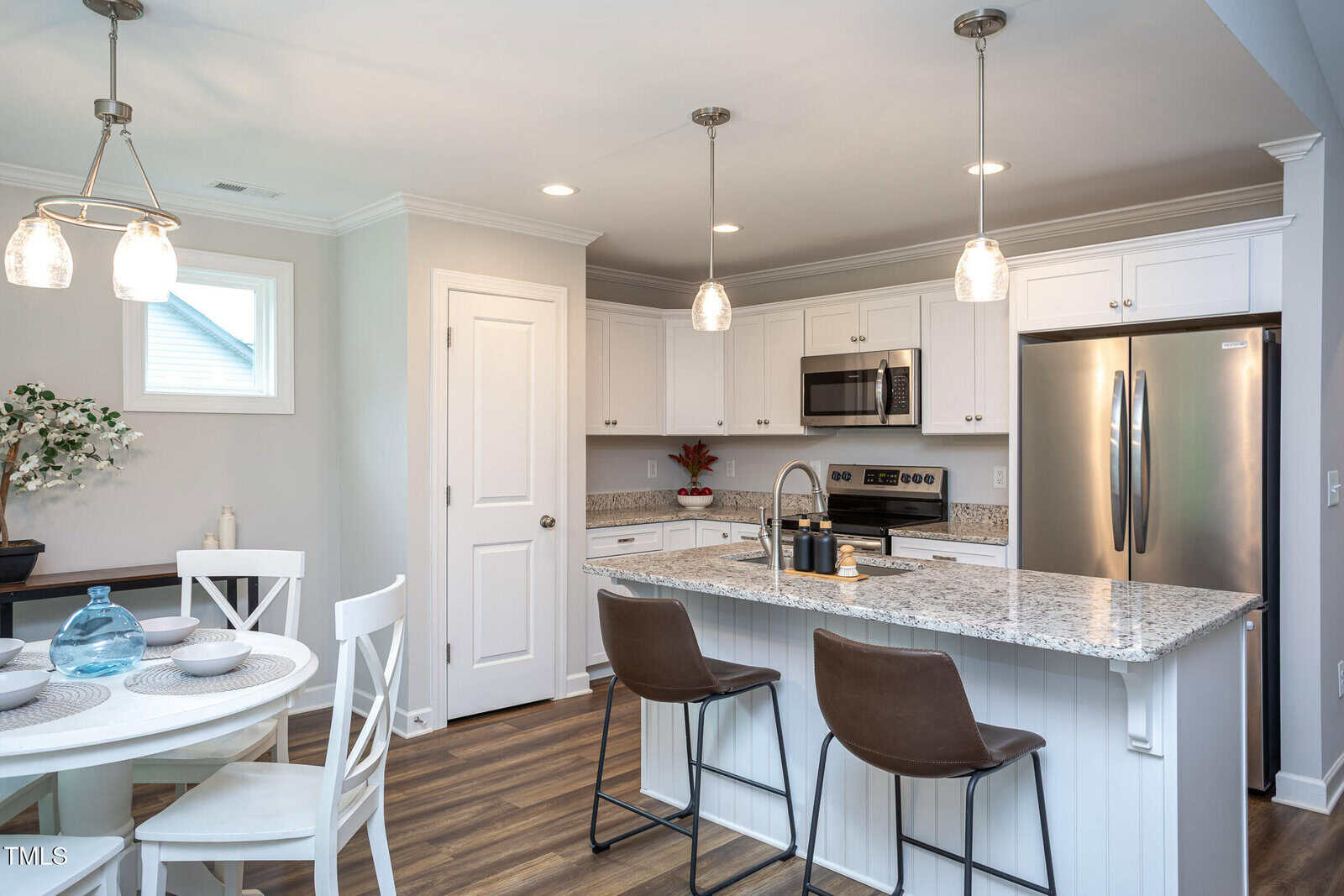 Modern kitchen with white cabinets, stainless steel appliances, granite countertops, pendant lights, and a center island with two brown chairs; dining table with four settings is visible in the corner.