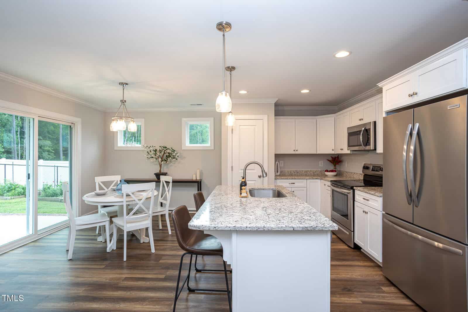 Modern kitchen with white cabinets, stainless steel appliances, granite countertops, a central island with two brown chairs, and a small dining table with four white chairs near large sliding glass doors.
