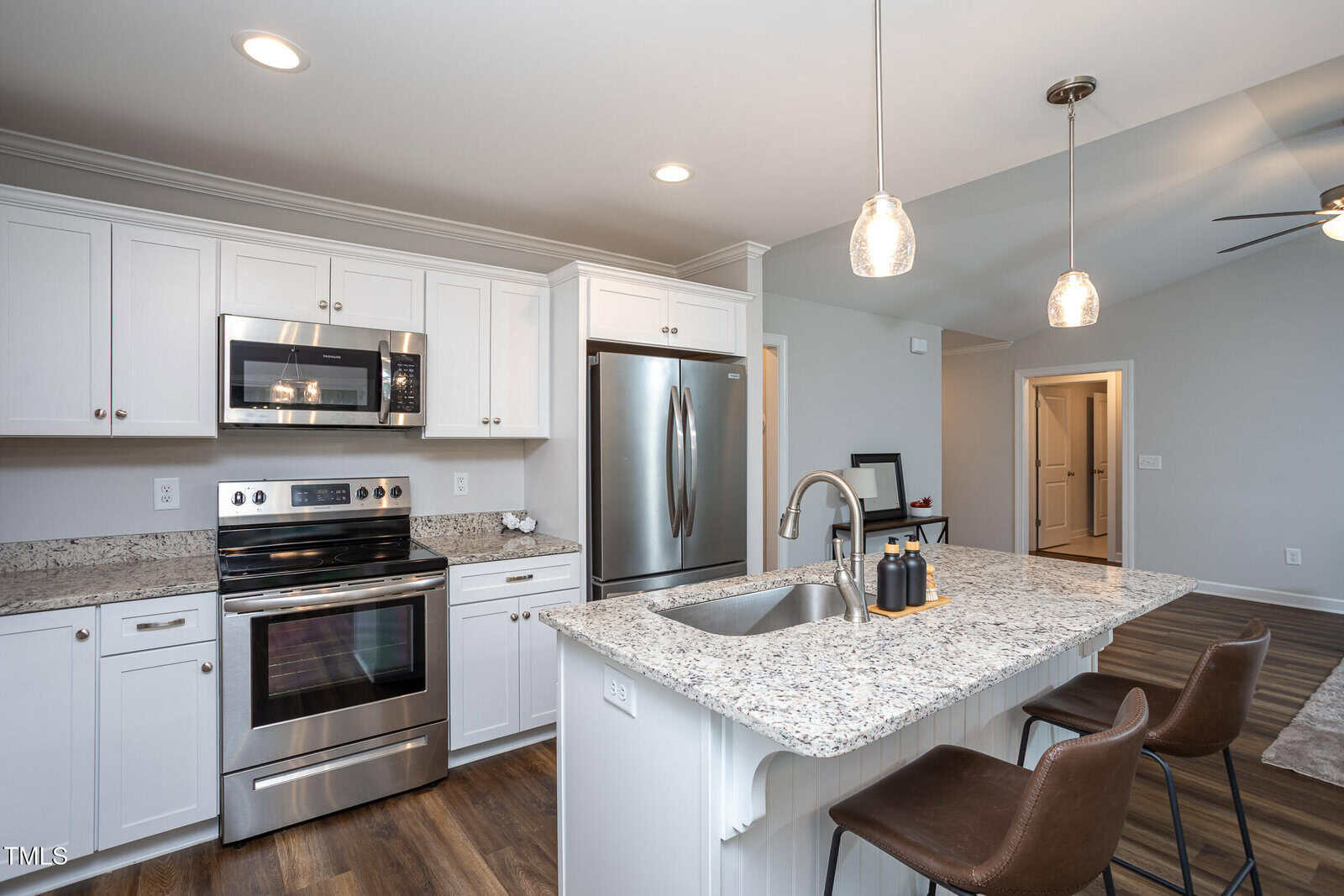 Modern kitchen with white cabinets, stainless steel appliances, a granite island with sink and two pendant lights, and two brown barstools on wood flooring. A hallway and ceiling fan are visible in the background.