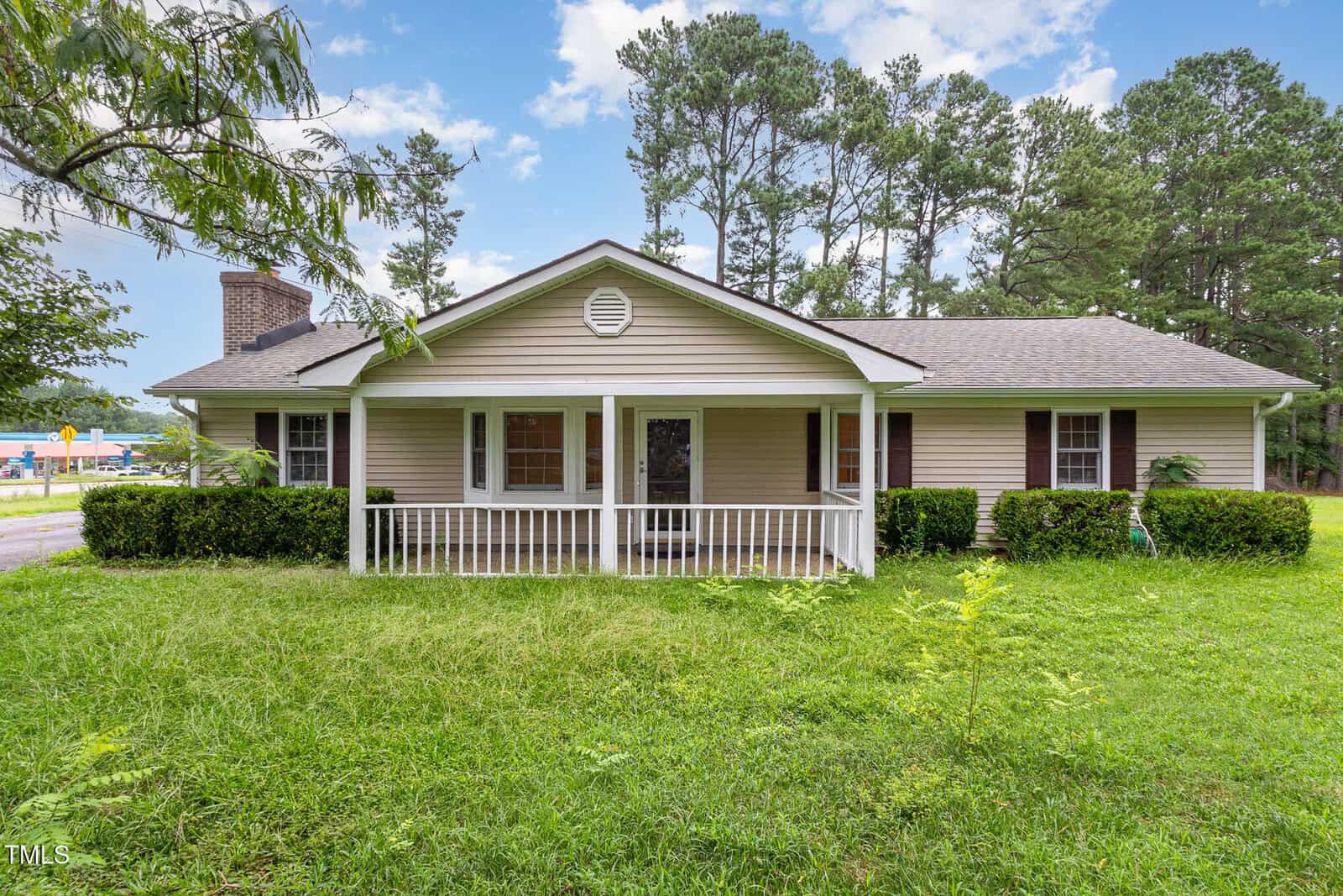 A single-story beige house with a small front porch and white railing, surrounded by green grass and bushes, and tall trees in the background under a partly cloudy sky.