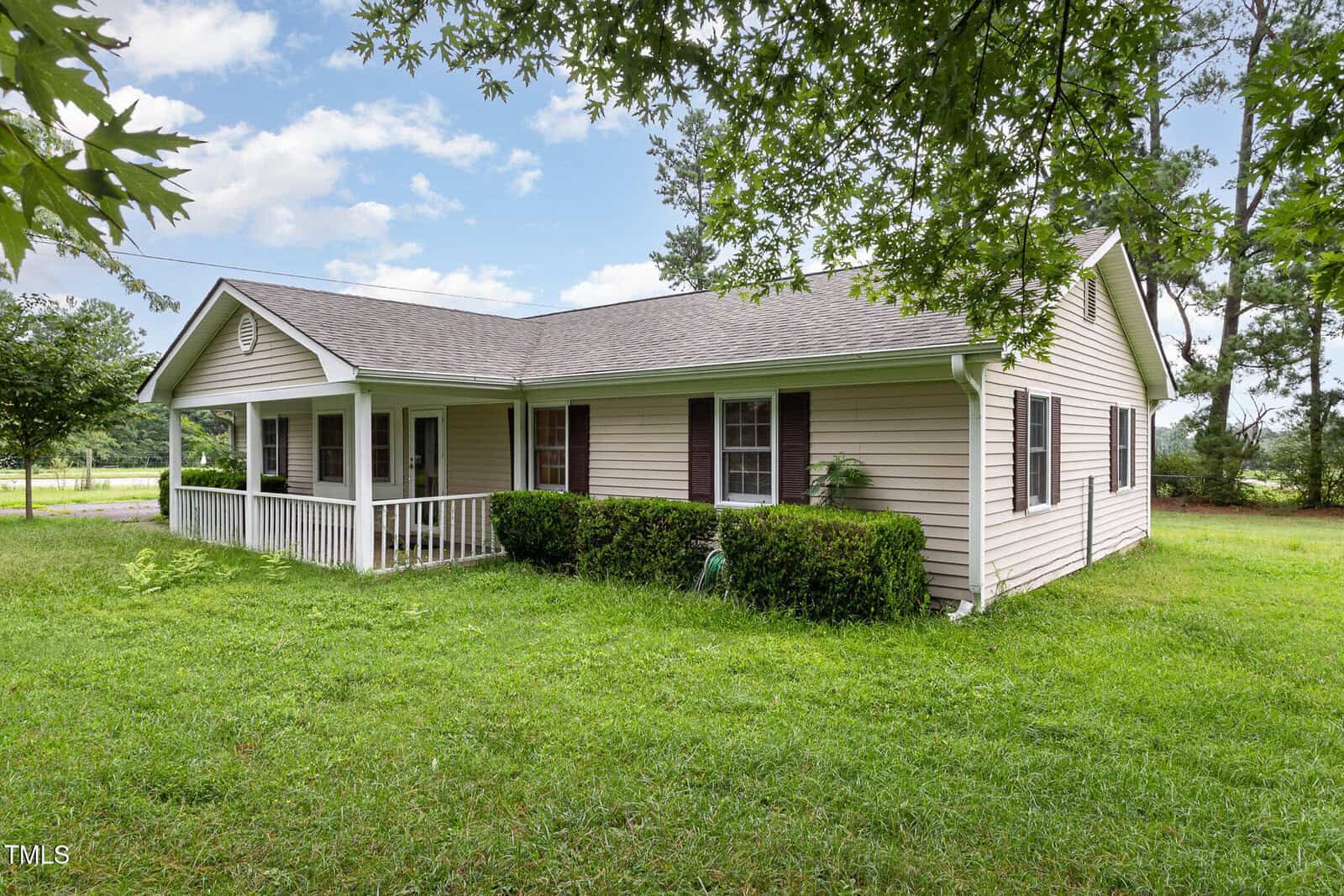 A single-story beige house with dark shutters, a covered front porch, and a well-kept lawn with shrubs, surrounded by green grass and trees under a partly cloudy sky.
