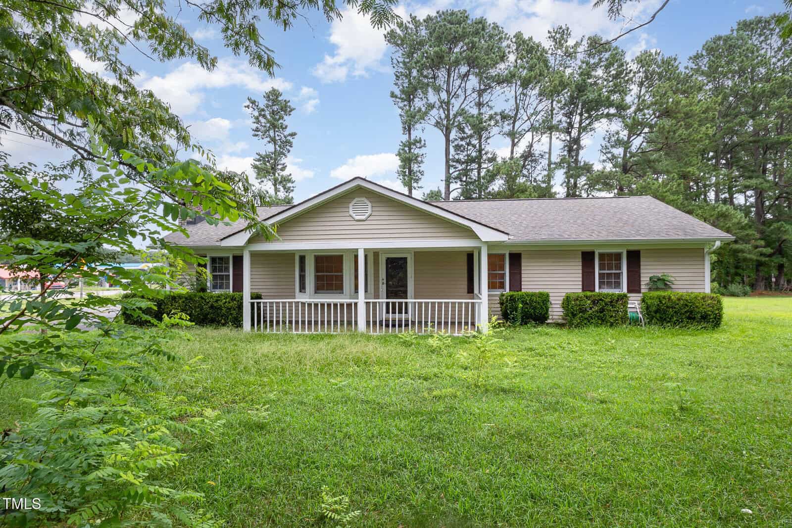 Single-story beige house with a gray roof, white trim, and front porch, surrounded by green grass and shrubs, with tall pine trees in the background under a partly cloudy sky.