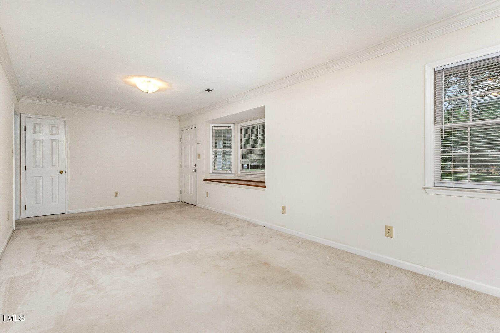 A spacious, empty room with off-white walls and carpet, two windows with blinds, a recessed wall shelf, and two closed white doors. Ceiling light fixture and baseboard trim are visible.