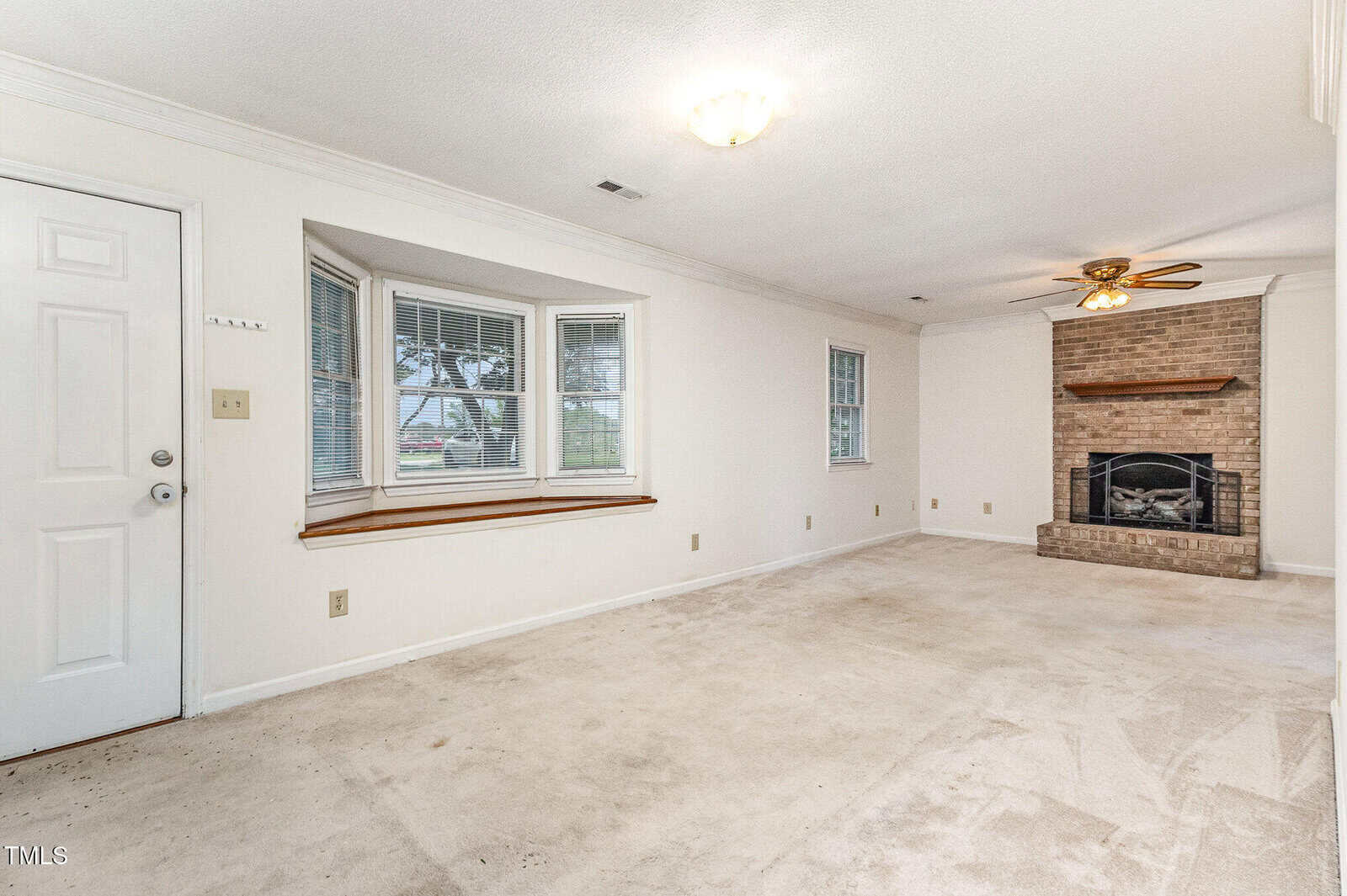 A spacious living room with white walls, beige carpet, a bay window with a wood ledge, a ceiling fan, and a brick fireplace. The room has minimal built-in features and a door on the left side.