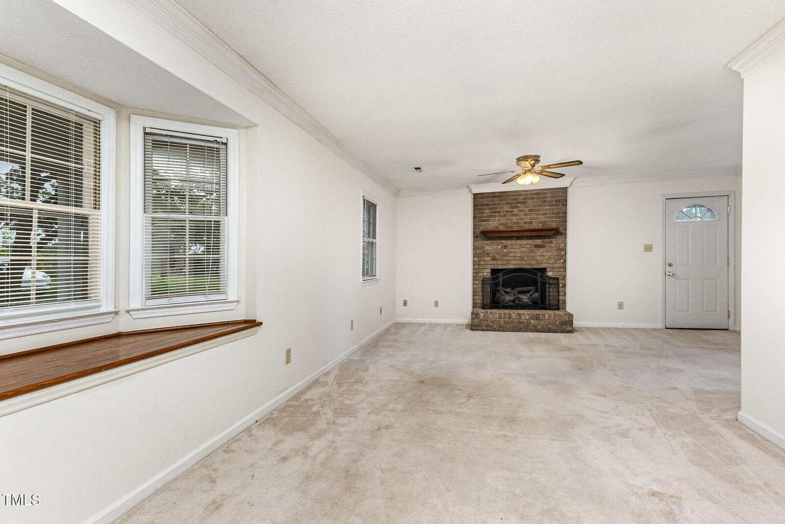 A carpeted living room with white walls, a ceiling fan, a brick fireplace, a white door, and two sets of windows including a bay window with a wooden ledge.