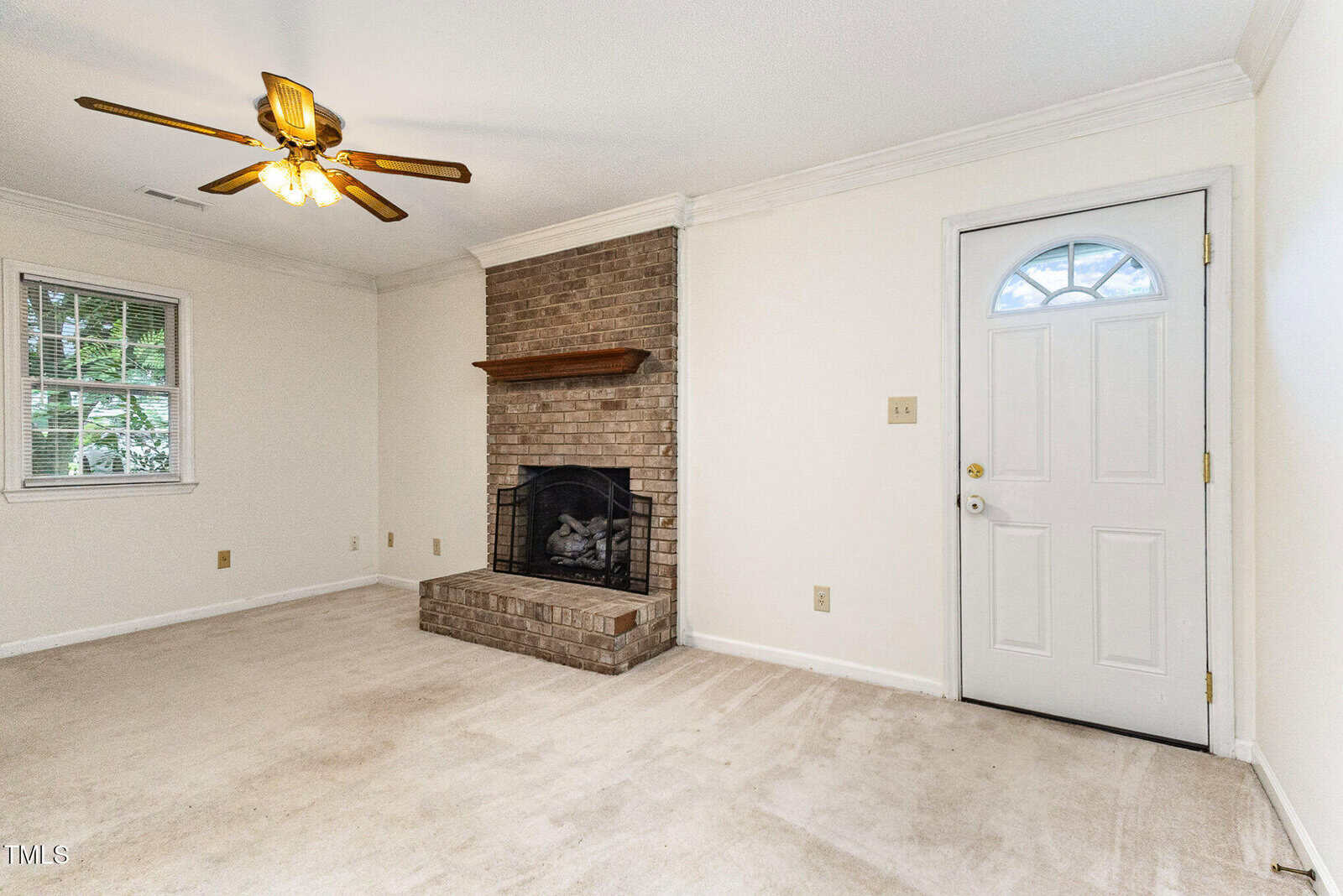 A carpeted room with a white door, window, and brick fireplace with a wooden mantel. A brass ceiling fan with lights hangs from the ceiling. Walls are painted off-white.