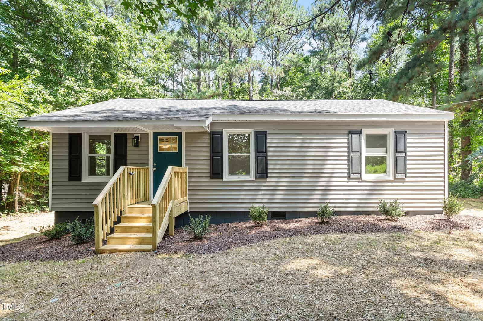 Single-story house with beige siding, black shutters, and a small wooden porch with stairs, surrounded by trees and mulch landscaping in the front yard on a sunny day.