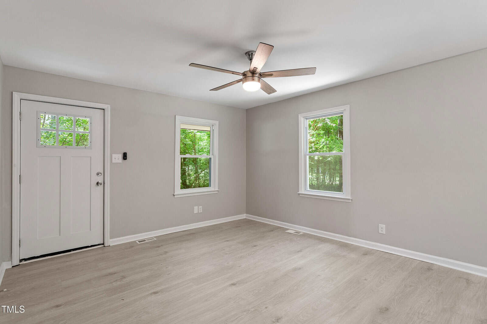 A bright, empty room with light wood floors, beige walls, a ceiling fan with light, two windows, and a white door. Green trees are visible through the windows.