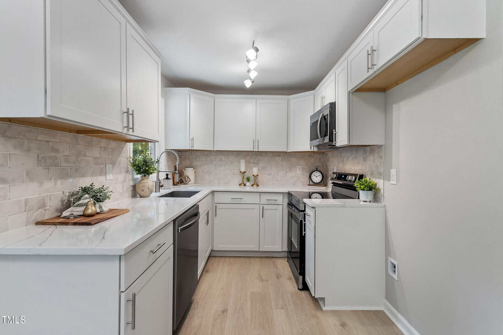 Modern kitchen with white cabinets, stainless steel appliances, light wood flooring, marble countertops, and a neutral tile backsplash. Decorative plants and kitchen items are neatly arranged on the counters.