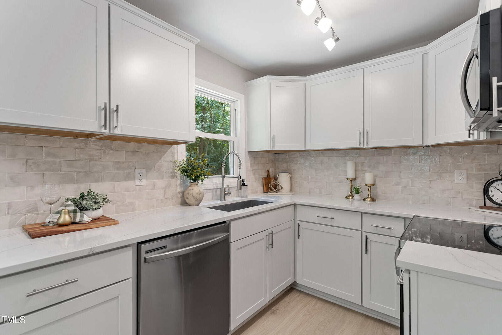 Modern kitchen with white cabinets, stainless steel appliances, a double-basin sink under a window, light-colored tile backsplash, and minimalist decor on the counters, including flowers and candles.