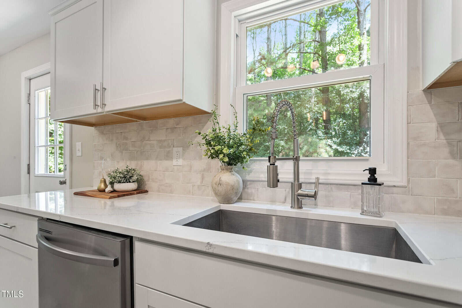 Modern kitchen with a white countertop, stainless steel sink, silver faucet, white cabinets, and a window with a view of greenery. A soap dispenser and vase with flowers sit next to the sink.