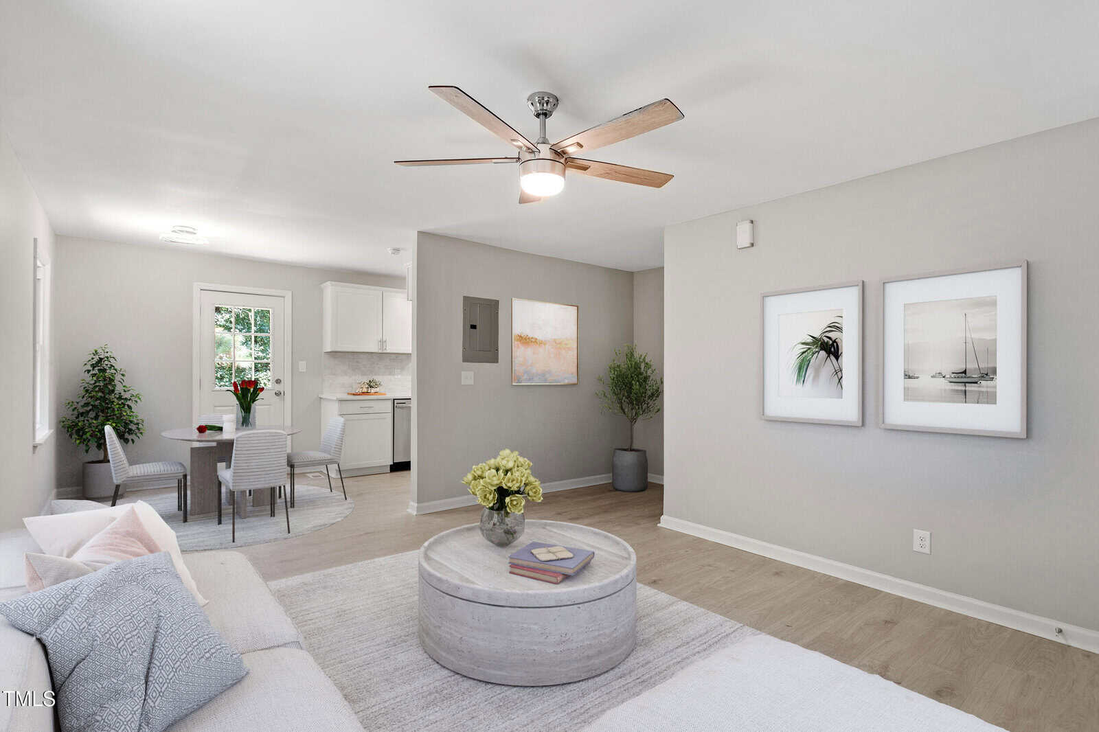 A bright, modern living room with light walls, wood floors, a ceiling fan, and neutral furniture. The space includes a round coffee table, two framed photos, and a view into a small dining area and kitchen.