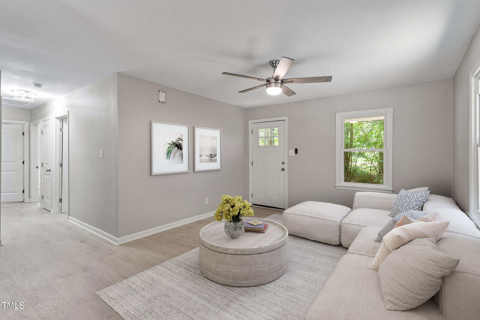 A bright, modern living room with light gray walls, wood floors, a round coffee table with a flower arrangement, a large sectional sofa with pillows, wall art, and a ceiling fan. Sunlight streams through the window.
