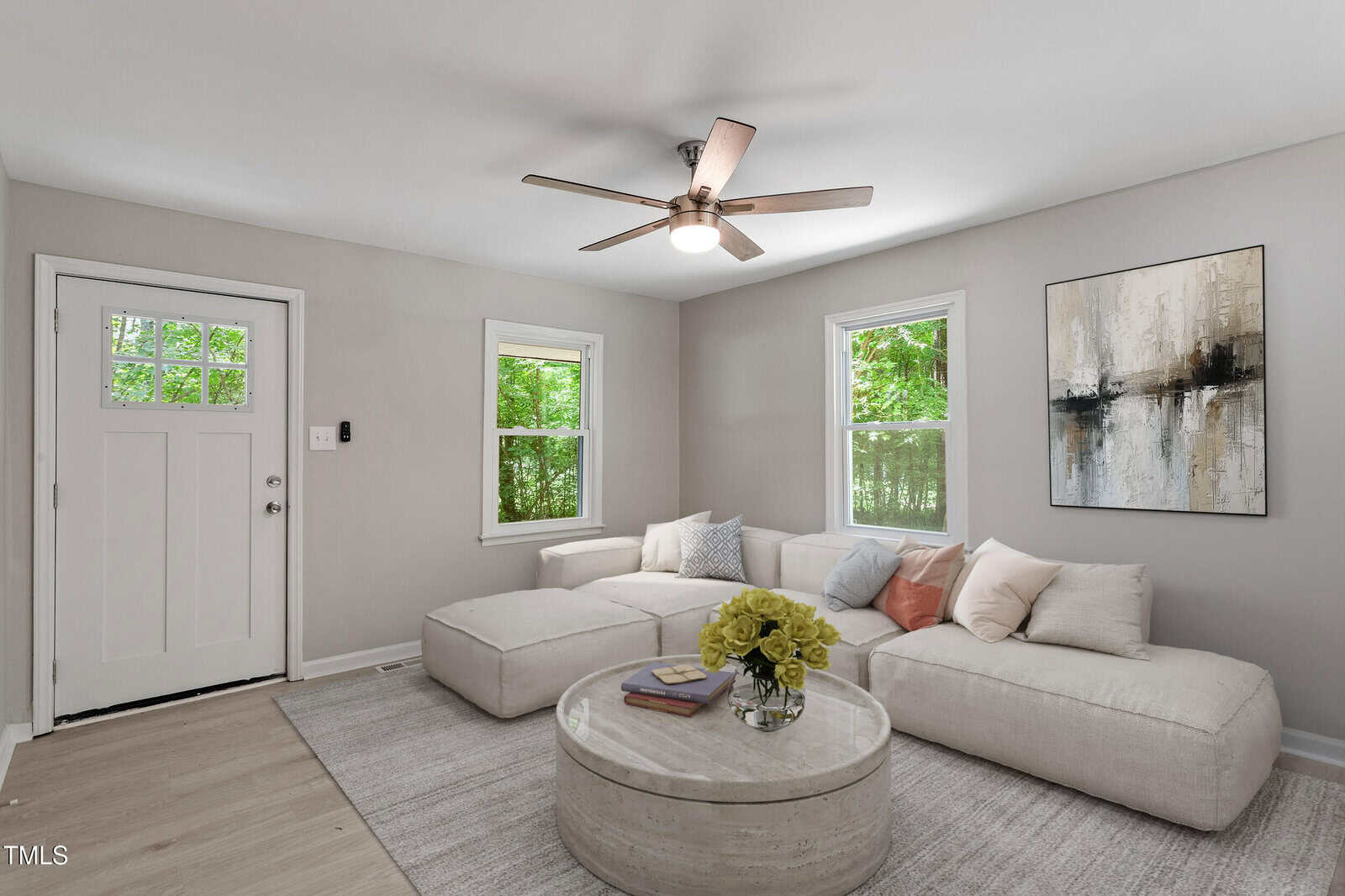 A modern living room with light gray walls, a beige sectional sofa with pillows, a round coffee table with flowers and books, abstract wall art, two windows, and a ceiling fan. Natural light fills the space.