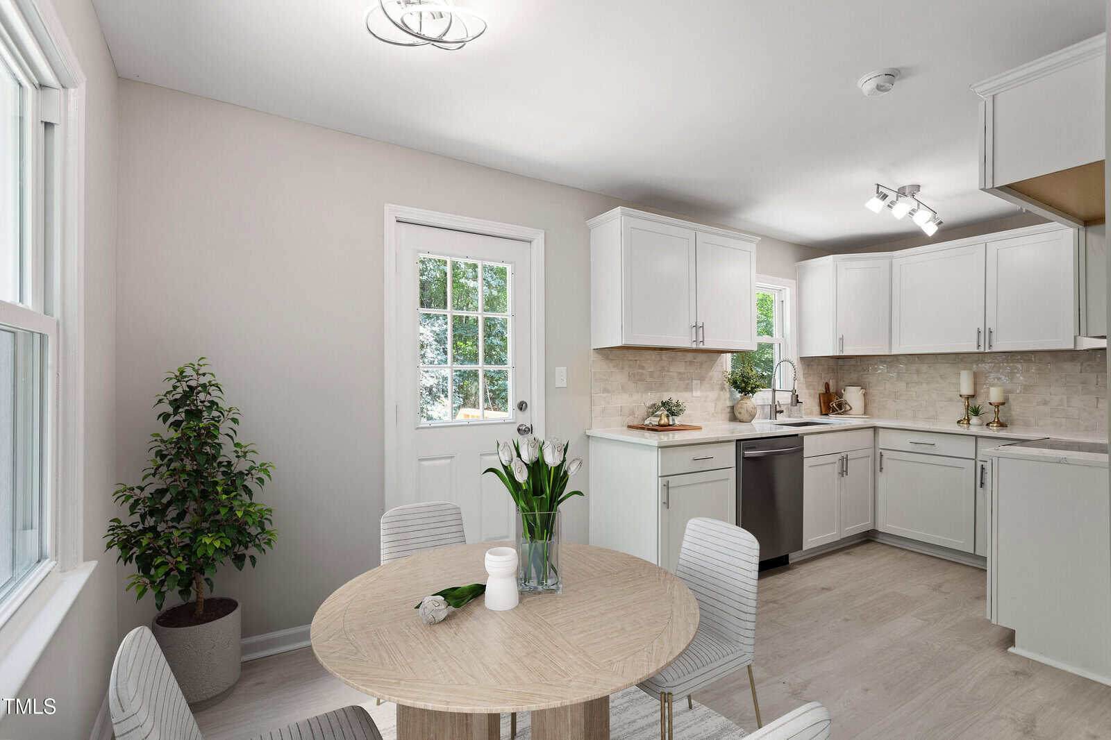 Bright, modern kitchen with white cabinets, light countertops, stainless steel appliances, and a round wooden dining table with four striped chairs. A vase of tulips and a potted plant add fresh touches to the space.