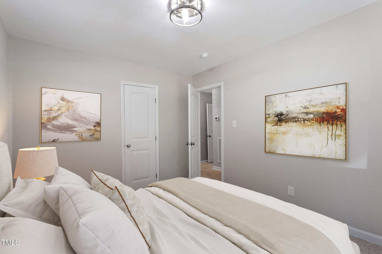 A modern bedroom with light gray walls, a bed with white and beige bedding, a bedside lamp, two abstract paintings on the walls, and a ceiling light fixture. The door is open, showing a hallway.