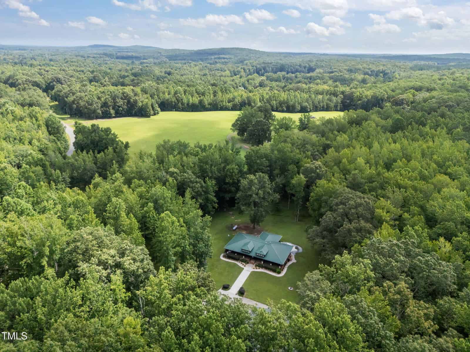 Aerial view of a green-roofed house surrounded by dense forest, with a large open grassy field and distant wooded hills under a partly cloudy sky. A winding road is visible through the trees.