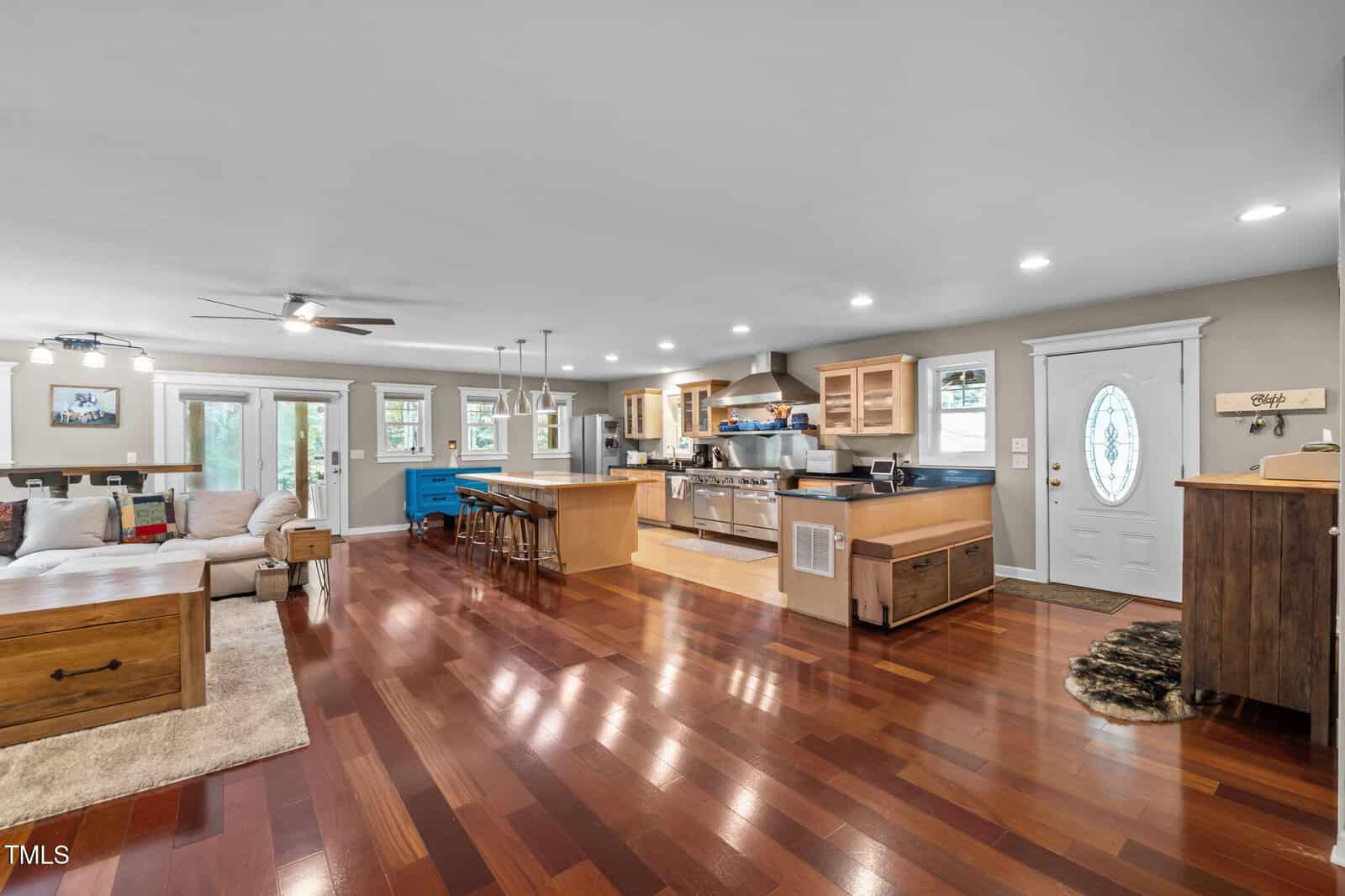 Open-concept living space with polished wood floors, a modern kitchen with stainless steel appliances, a central island with bar stools, a beige sectional sofa, and large windows for natural light.