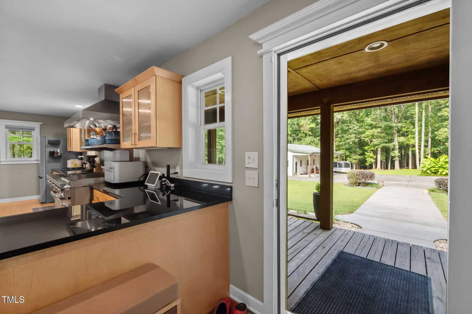 A view from a kitchen with light wood cabinets and black countertops, looking out through an open door onto a covered porch and yard with trees and a paved driveway.