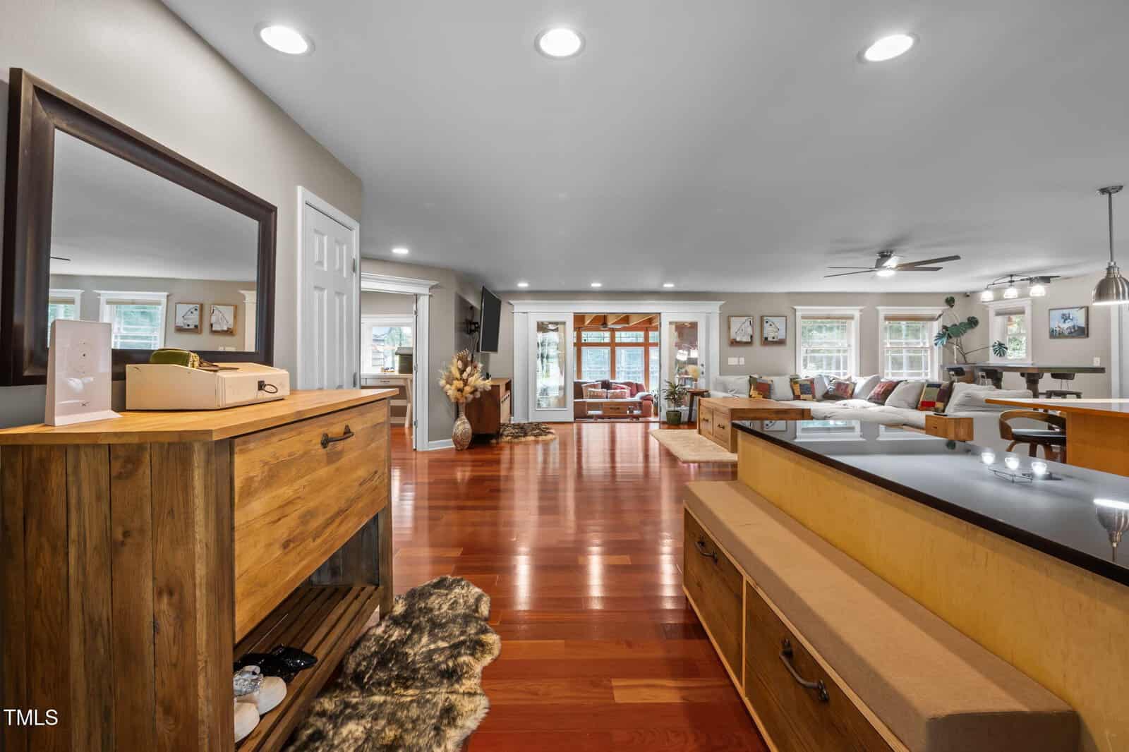Spacious open-concept living area with wood floors, recessed lighting, and large windows. A wooden entry table stands on the left, and a living room with a sectional sofa is visible in the background.