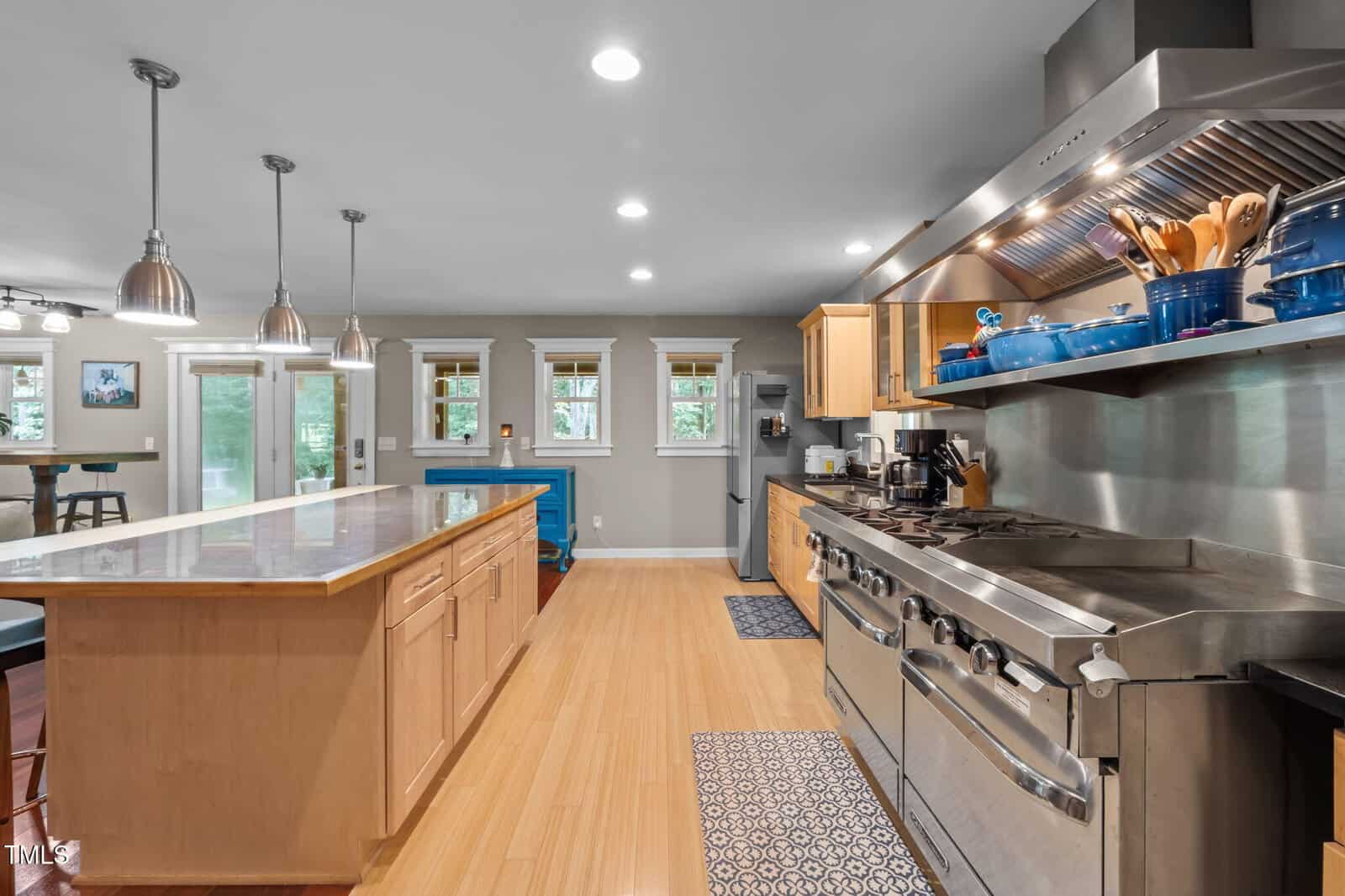 Spacious kitchen with light wood cabinets, large center island, stainless steel appliances, gas range, blue cookware, and hardwood floors; pendant lights and windows provide ample light.