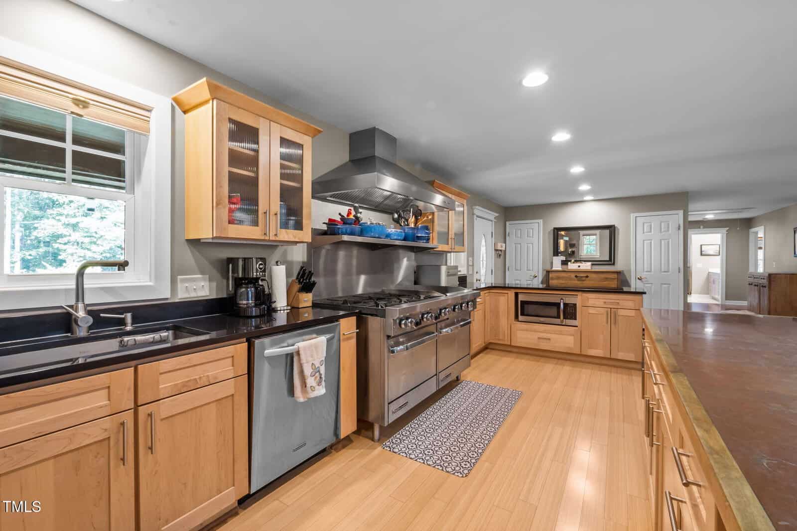 Spacious kitchen with light wood cabinets, stainless steel appliances, a large stove, black countertops, double sink, and a window above the sink. The floor is light wood, and recessed lighting brightens the room.