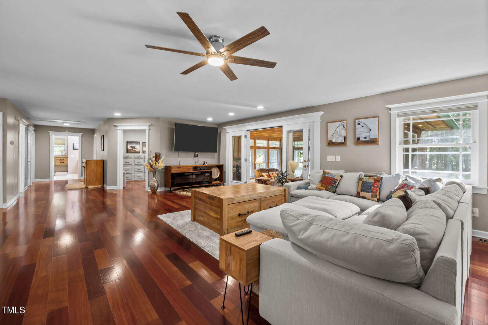 Spacious living room with wood floors, a large sectional sofa, wooden coffee table, wall-mounted TV, ceiling fan, and windows letting in natural light; adjacent rooms visible in the background.