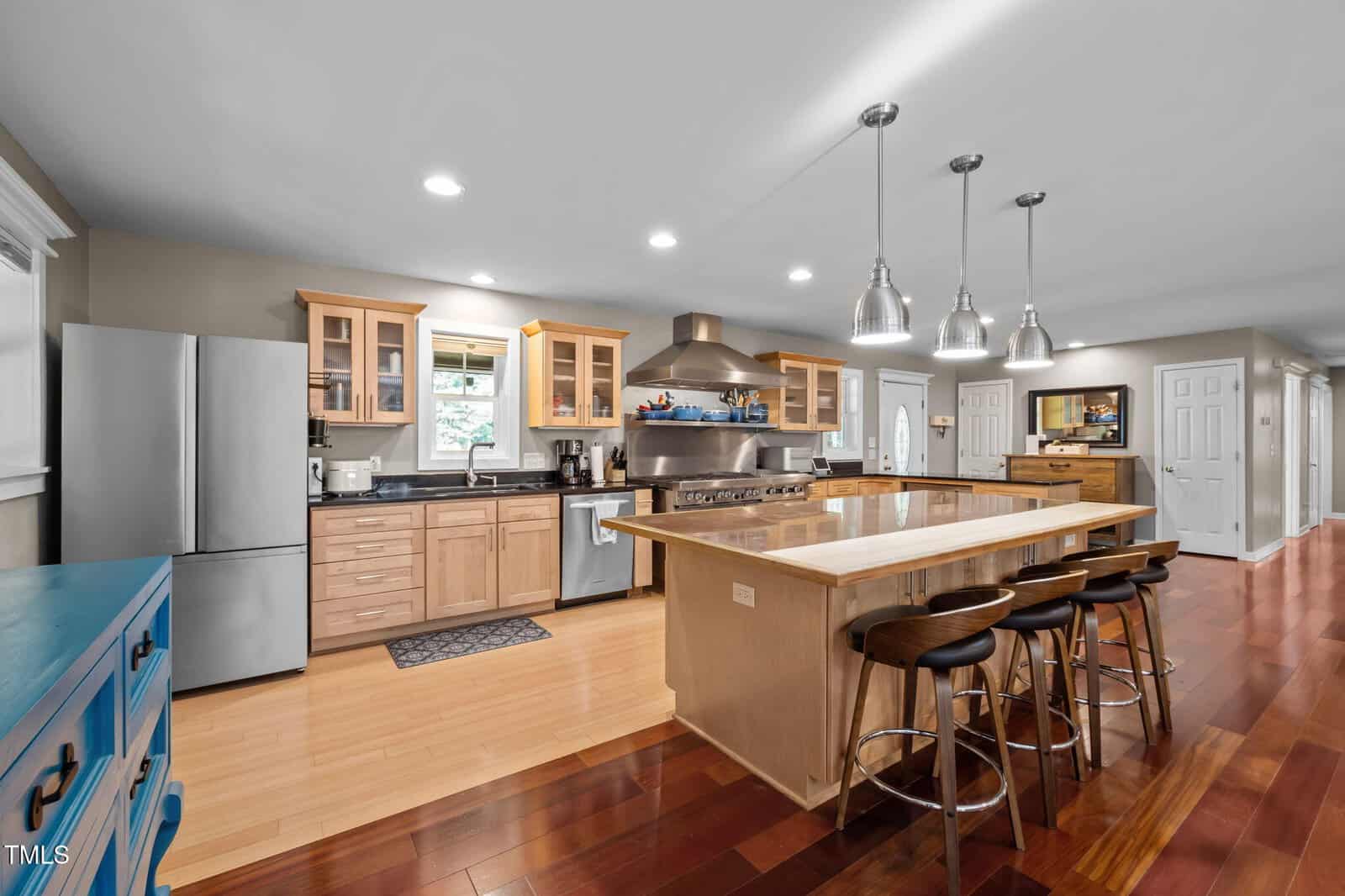 Spacious modern kitchen with stainless steel appliances, wooden cabinets, large island with stools, pendant lighting, and hardwood floors.