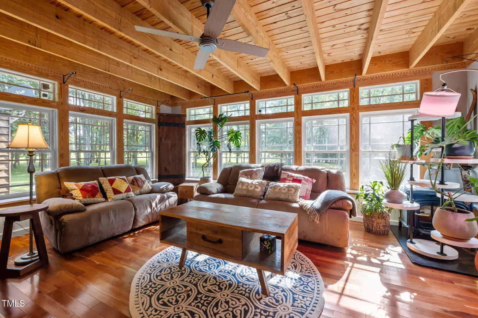 A sunroom with wooden ceiling beams, large windows, two sofas with patterned pillows, a wooden coffee table on a round rug, several potted plants, and a floor lamp, all bathed in natural light.