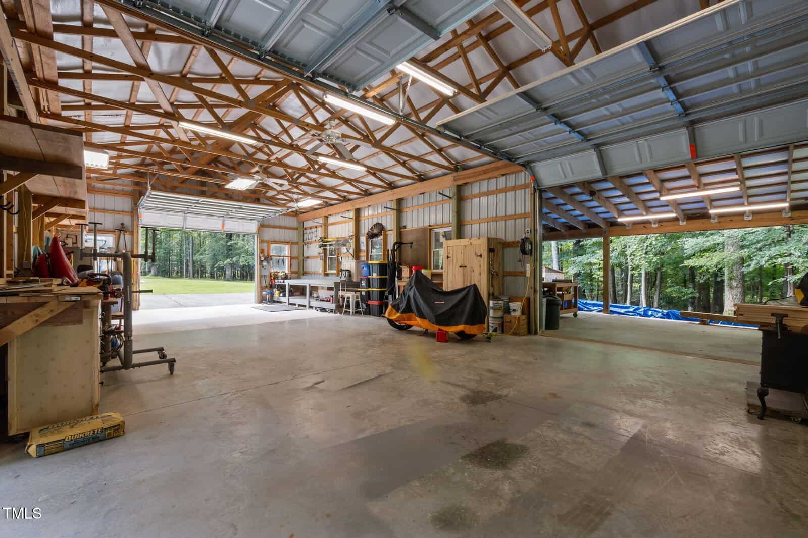 Spacious metal workshop with open garage doors, exposed beams, multiple workbenches, various tools, shelving, and covered equipment. Trees and grass are visible outside through the open doors.