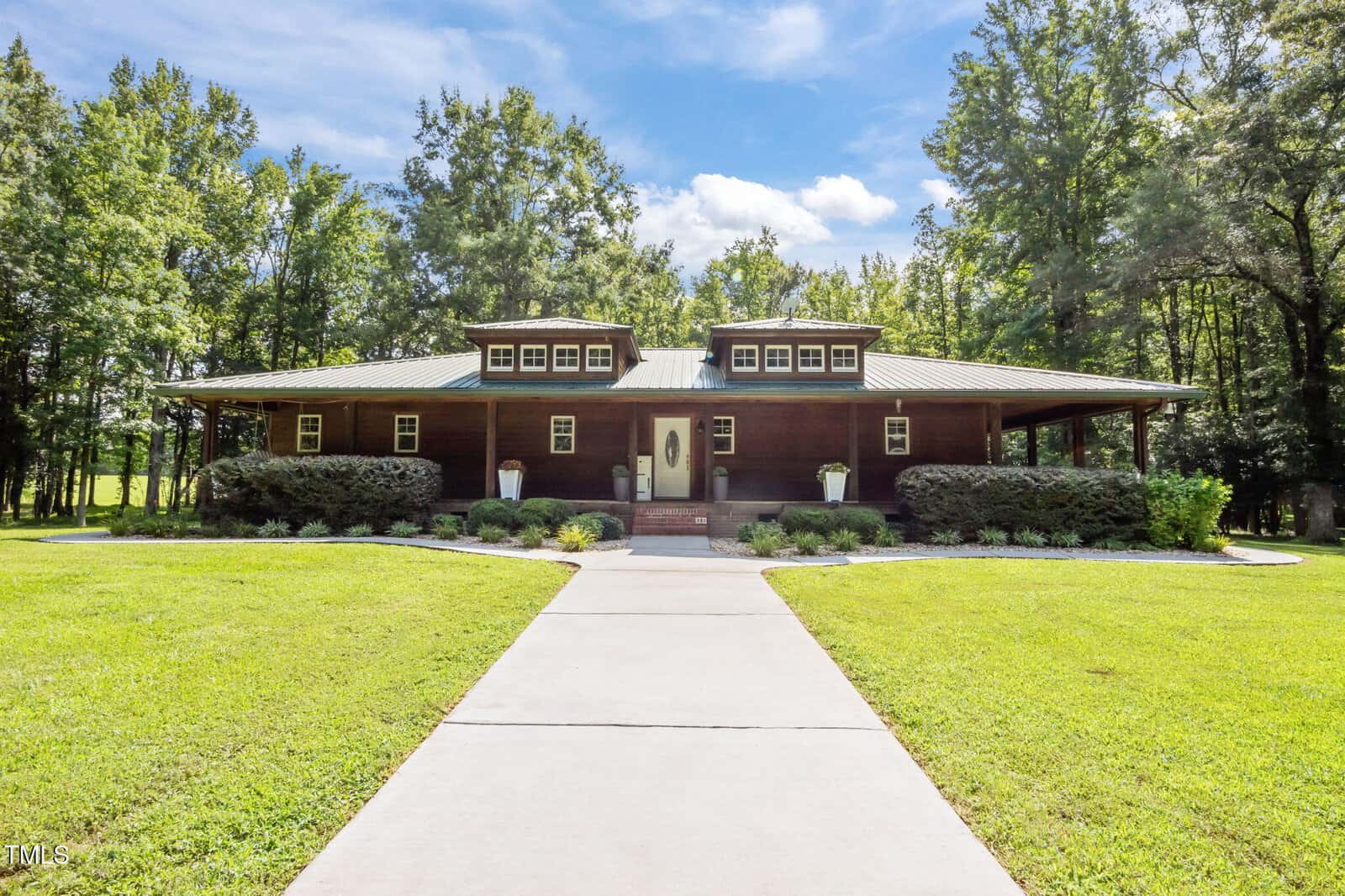A single-story wooden house with a metal roof at 4116 Blue Mountain Road, Oxford, featuring two dormer windows, a centered front door, and a wide wraparound porch. Surrounded by trees and landscaped bushes with a concrete walkway to the entrance.