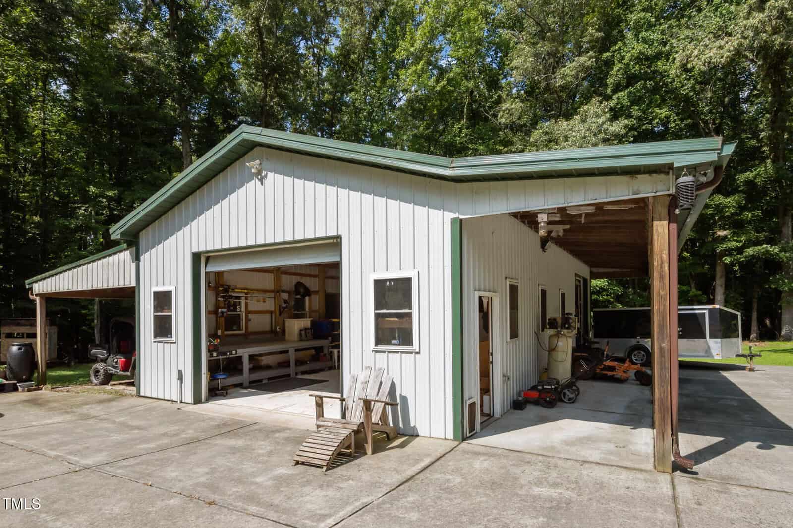 A metal garage building with an open garage door showing tools and workbenches inside. There are two wooden chairs outside and a covered area on the right. The building is surrounded by trees and a concrete driveway.