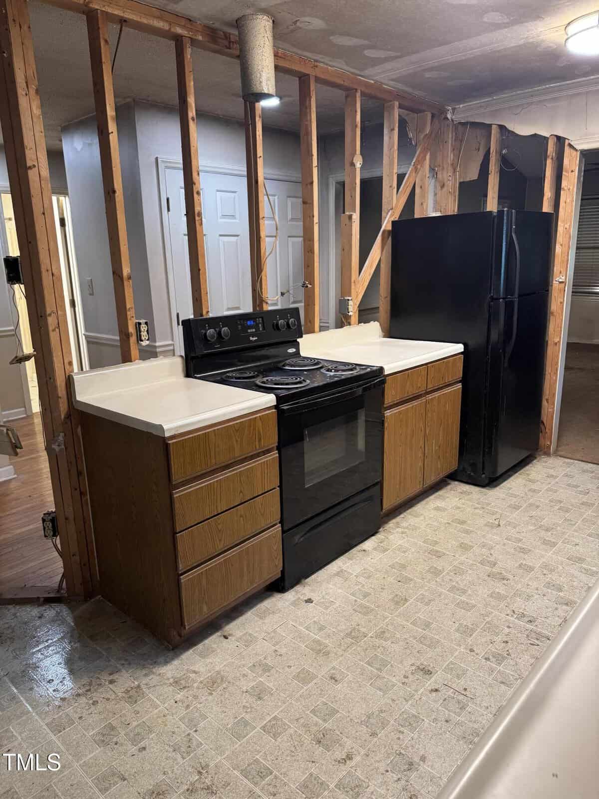 A partially renovated kitchen with exposed wall studs, a black stove and refrigerator, brown cabinets with white countertops, and a tiled floor. The ceiling and lighting appear unfinished.