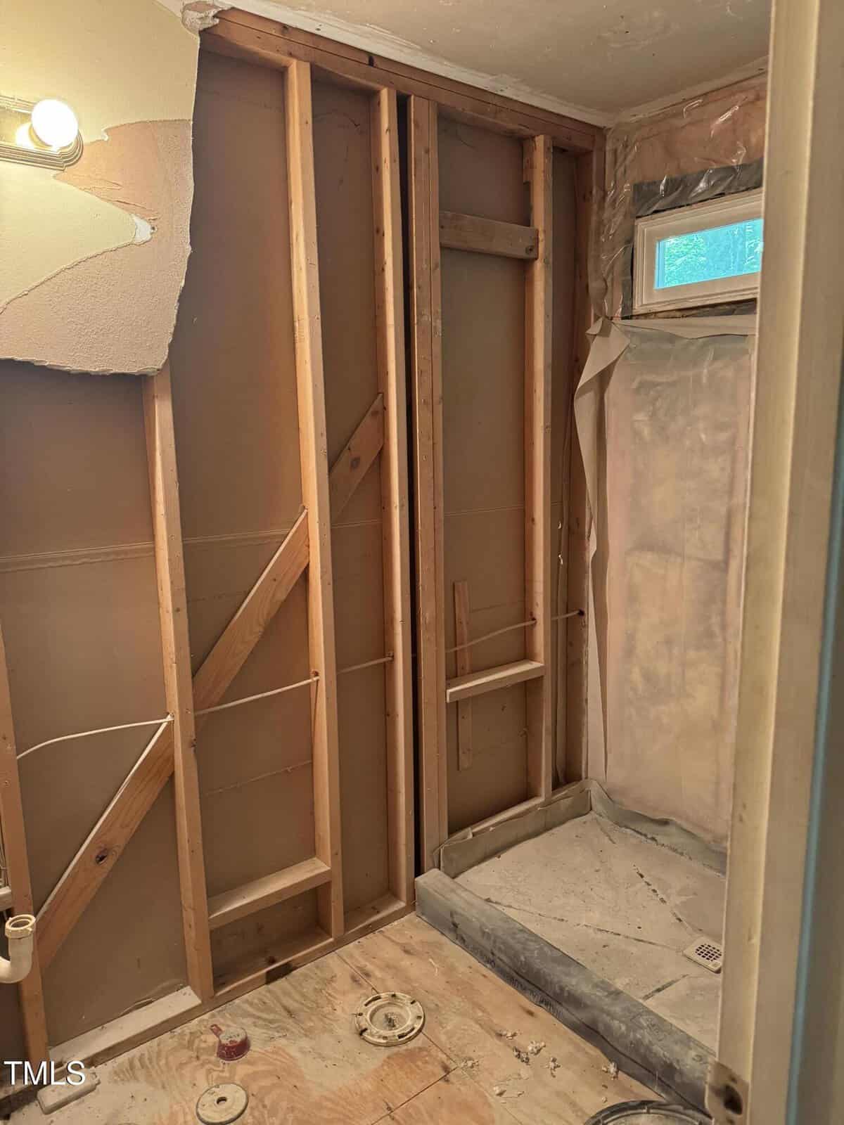 Bathroom under renovation with exposed wall studs, partial drywall removed, plumbing visible, and a shower area with no tiles. A small window is above the shower. A light fixture is visible on the upper left wall.