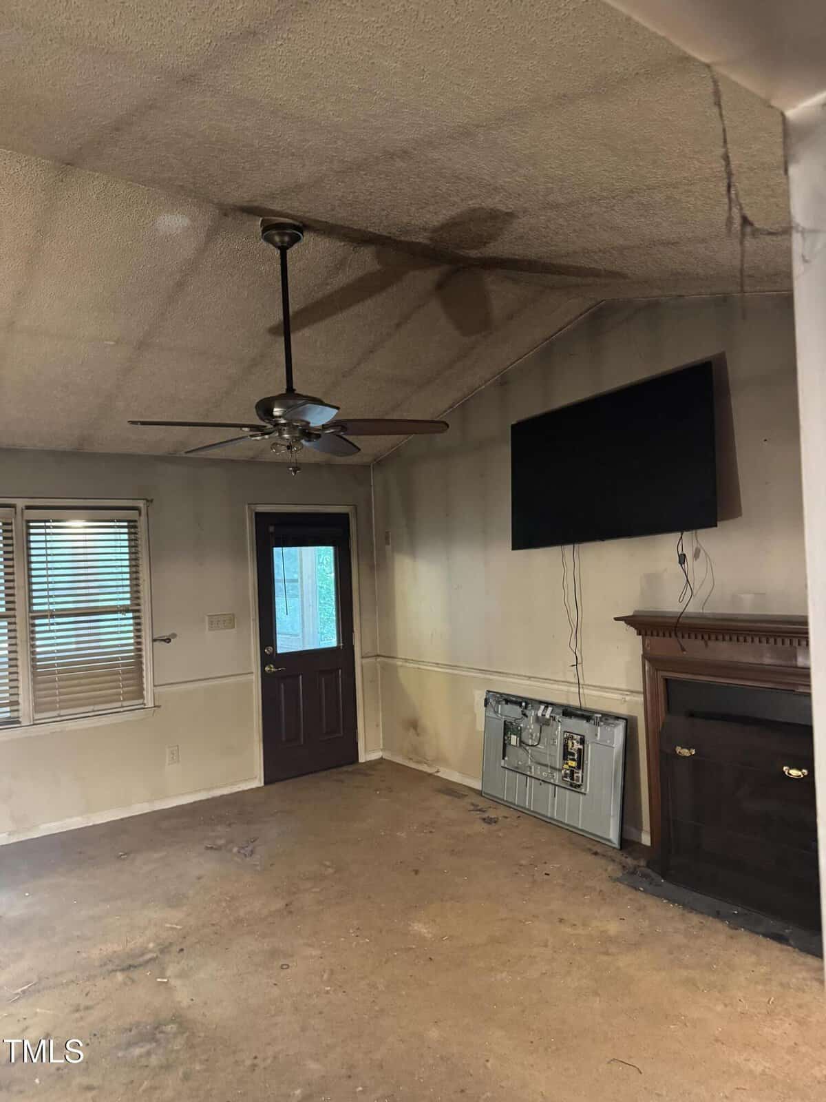A dimly lit, empty room with stained walls and ceiling, dirty carpet, and visible grime. There's a ceiling fan, a wall-mounted TV, a fireplace, a dark door, and windows with blinds. Cables hang below the TV.