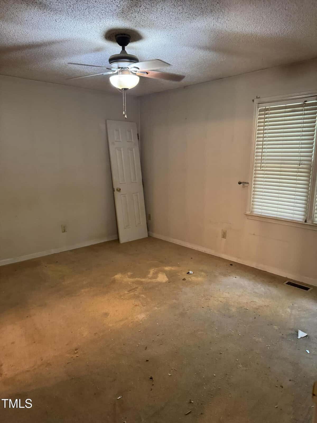 An empty room with stained carpet, a ceiling fan with light, a window with blinds, and a detached door propped against the wall. The walls are plain, and there is visible dirt and debris on the floor.