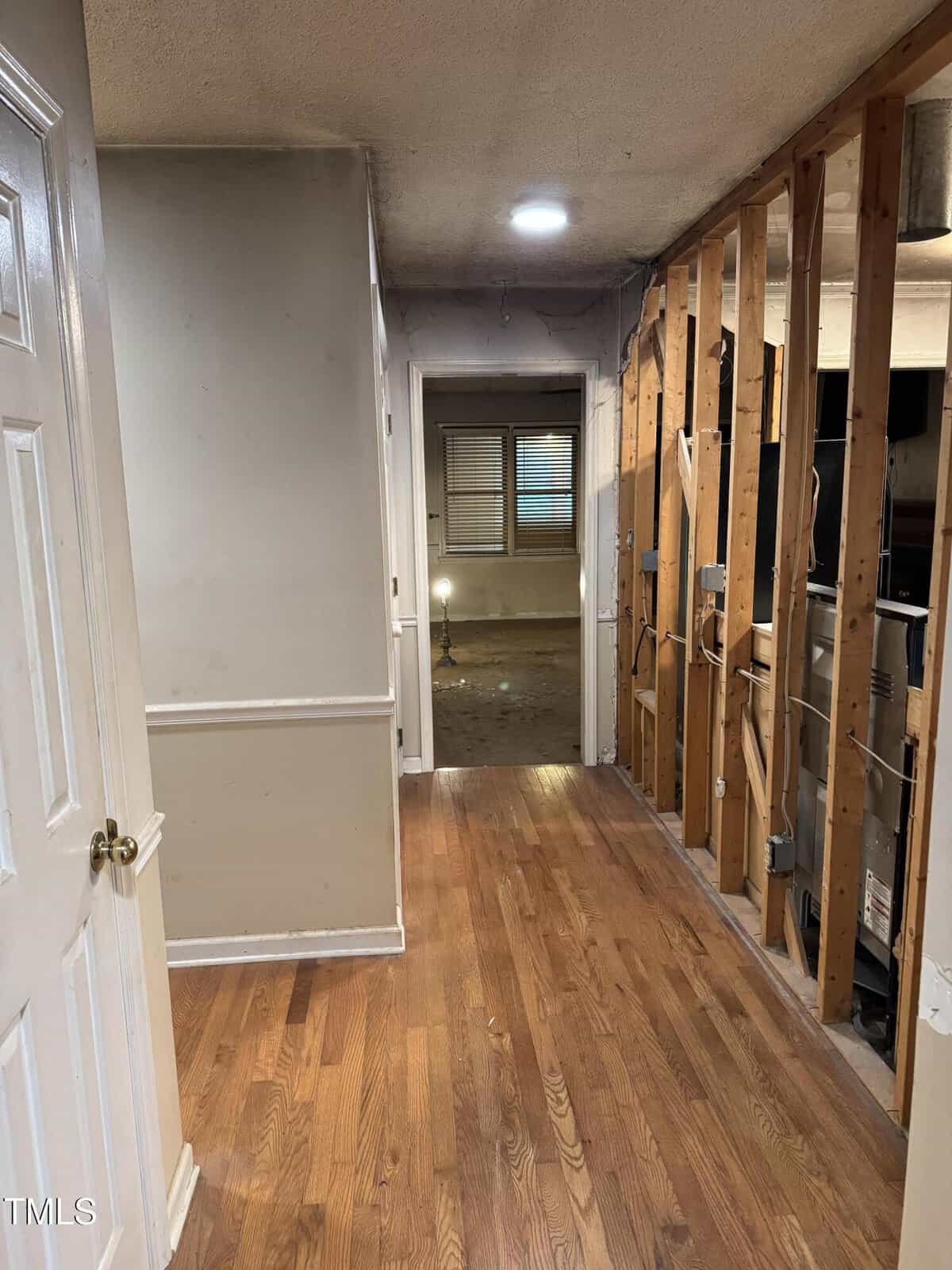 A hallway with hardwood floors leads to a room with a lit lamp. The right wall is partially demolished, exposing wooden studs and pipes, while the left wall is intact and painted white.