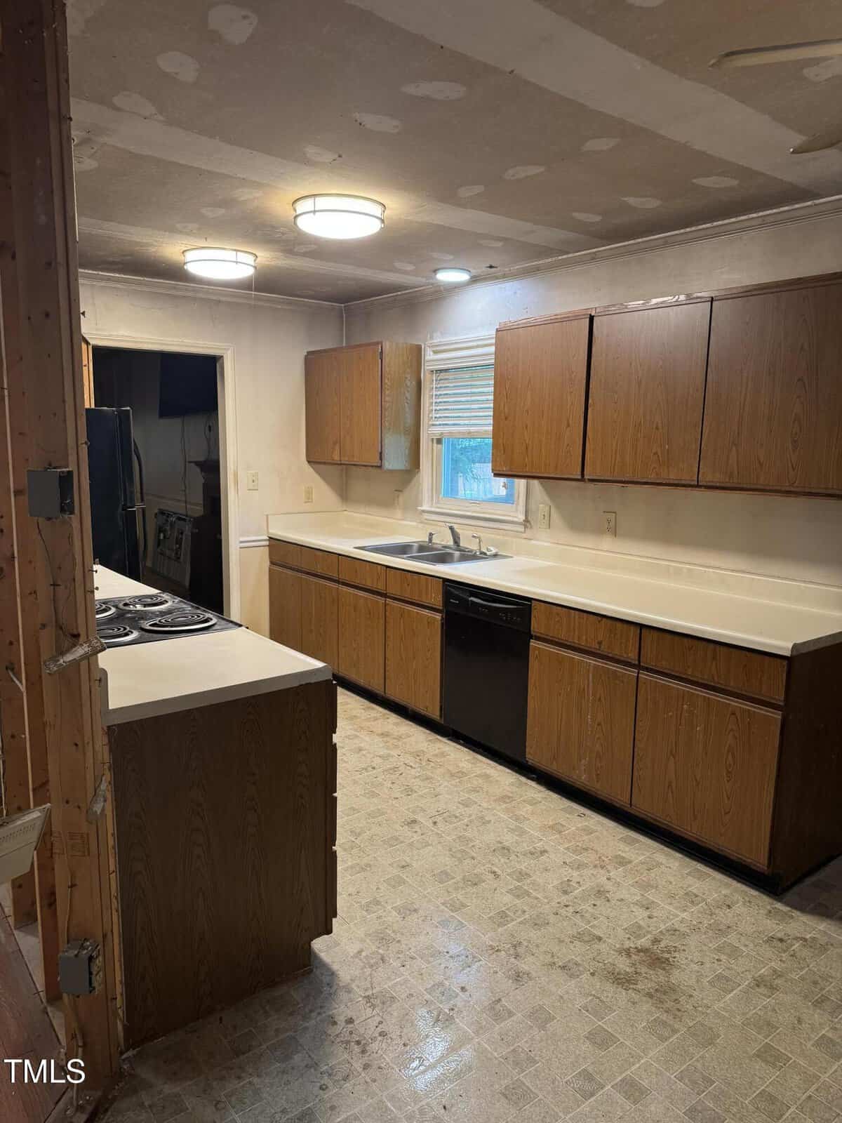 A kitchen with unfinished ceilings, wood cabinets, a white countertop, a dishwasher, stove, and a window above the sink; the floor has light-colored, worn tiles.