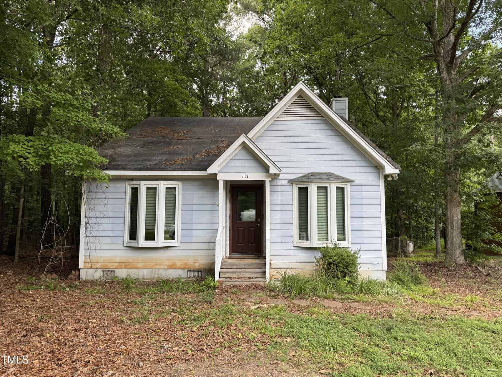 A small, light blue house with white trim and a dark roof stands in front of tall trees. The house has a central front door, two bay windows, and a patchy yard with sparse grass and fallen leaves.