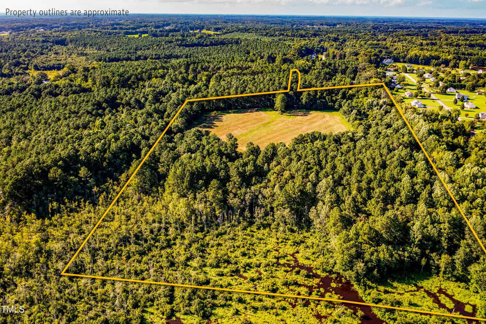 Aerial view of a large, mostly wooded property outlined in yellow, with a cleared field in the center and neighboring homes and forest surrounding the area. Text notes property outlines are approximate.